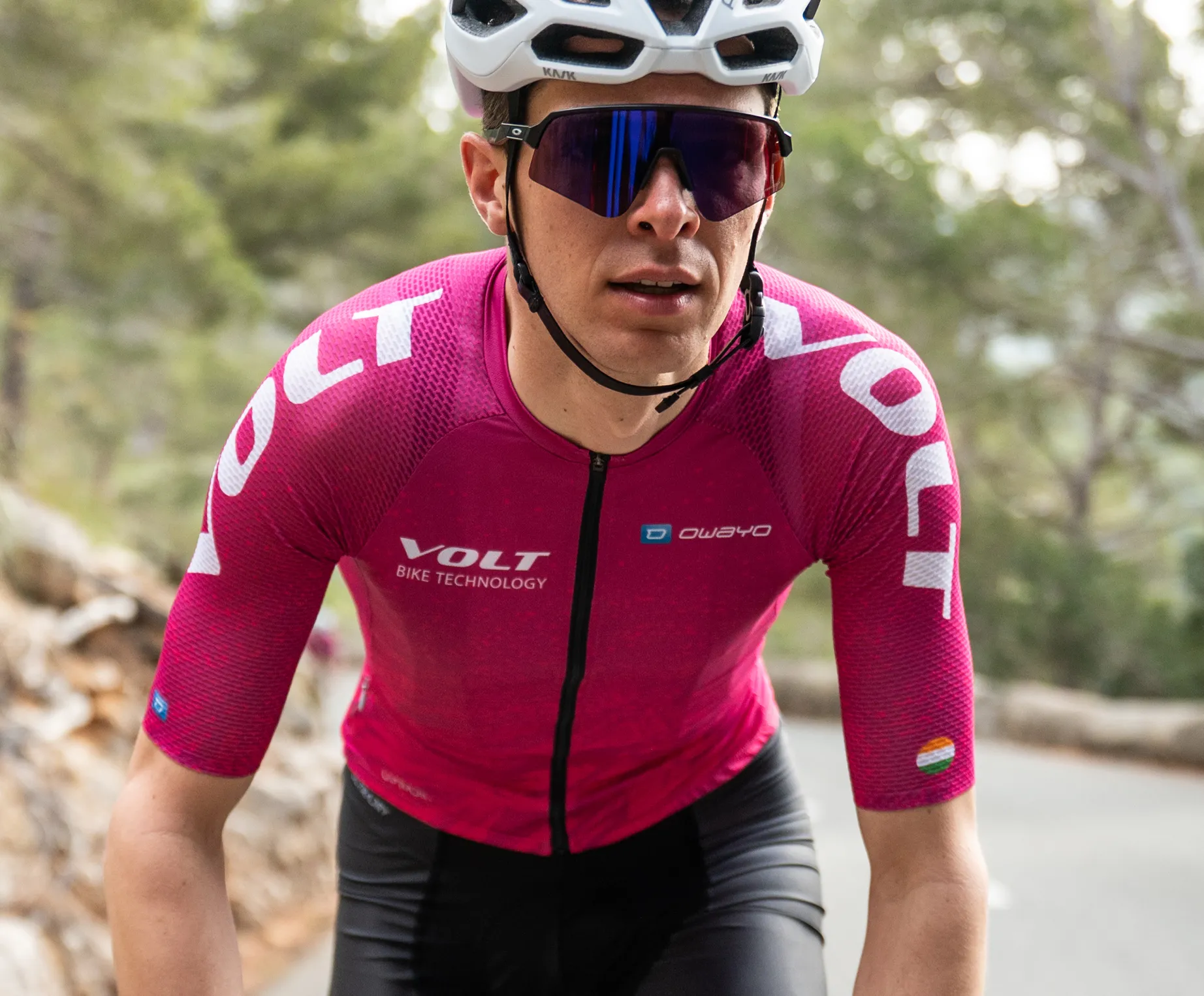 Ein Radfahrer in einem pinken, ärmellosen Trikot mit dem Logo 'VOLT BIKE TECHNOLOGY' und dem 'owayo'-Logo auf der Brust, der auf einer Straße in einem bewaldeten Gebiet fährt.