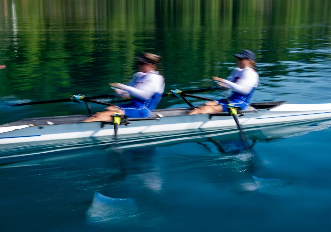 Zwei Ruderer in blauer Sportkleidung rudern synchron auf einem ruhigen Gewässer.