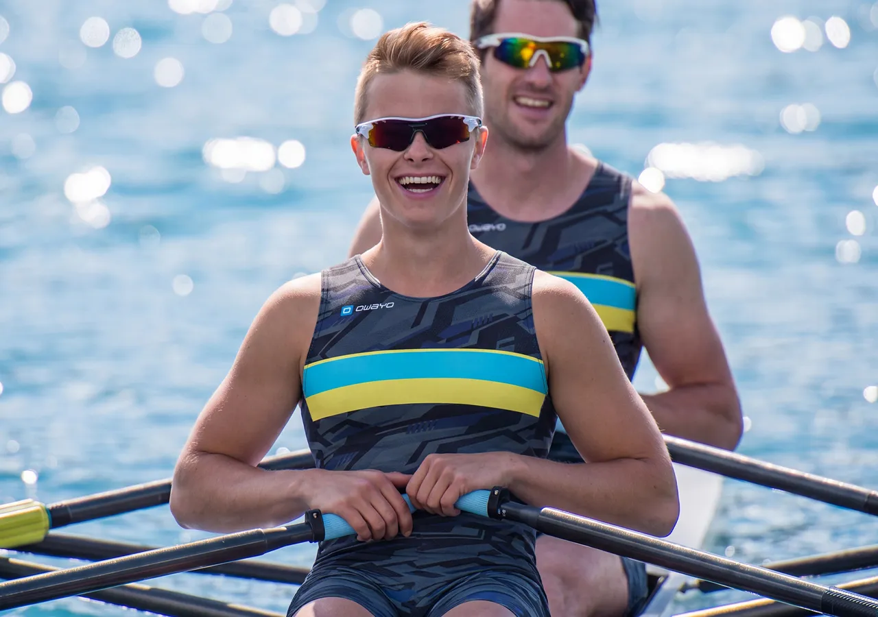 Zwei Athleten in stylischer Sportbekleidung sitzen in einem Ruderboot und lächeln in die Kamera, während sie auf einem funkelnden Wasser paddeln.