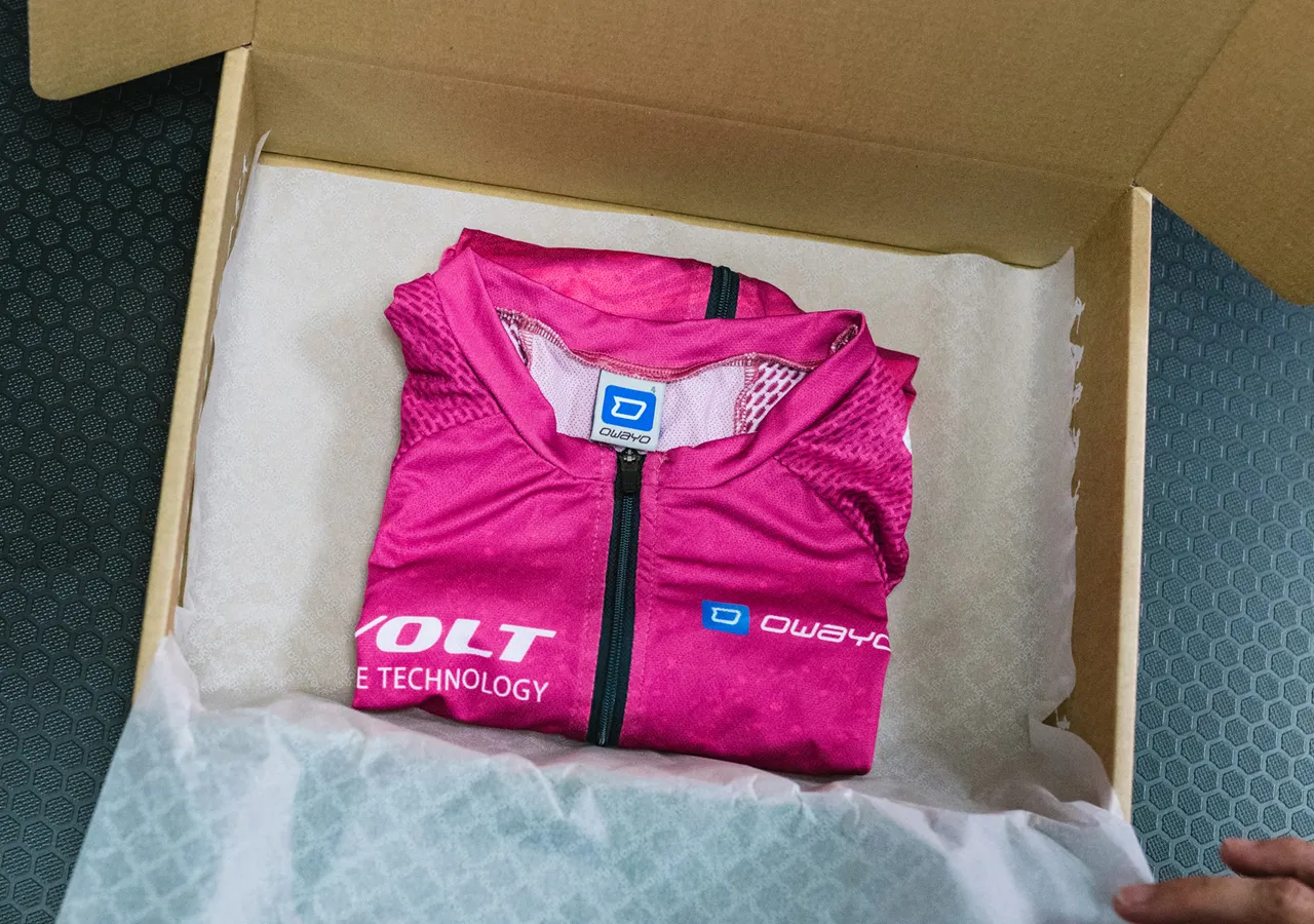 Ein gefaltetes pinkes, ärmelloses Sporttrikot mit dem Logo von owayo liegt in einer Pappschachtel auf weißem Papier.