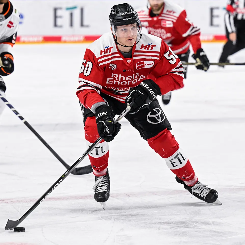 Ein Eishockeyspieler in einem roten Trikot mit der Nummer 50 und dem Schriftzug 'Rhein Energie' auf dem Eis, der einen Puck mit seinem Schläger kontrolliert.