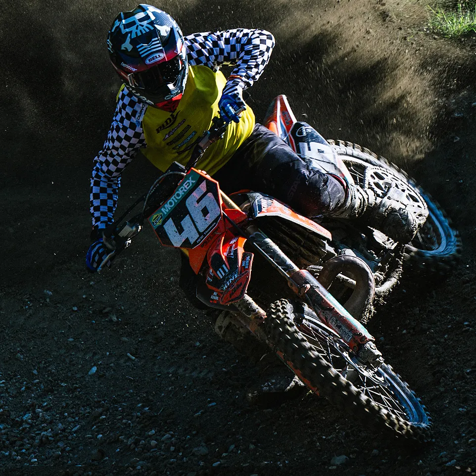 Ein Motocross-Fahrer in einer gelben Jacke mit schwarz-weiß kariertem Ärmel und einem Helm fährt eine orangefarbene Motorrad auf einer staubigen Strecke.