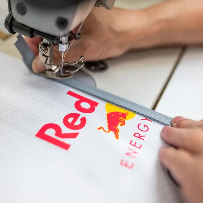 Eine Nahaufnahme einer Nähmaschine, die ein weißes Sportbekleidungsstück mit dem Red Bull-Logo näht, während eine Person das Material hält.