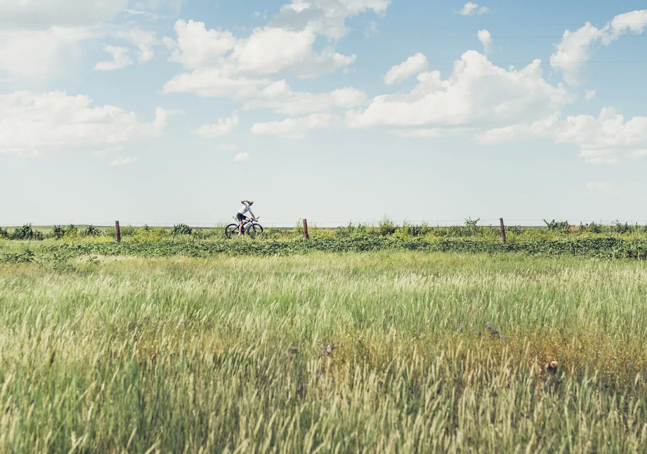 Eine Person fährt auf einem Fahrrad durch ein grünes Feld unter einem blauen Himmel mit weißen Wolken.