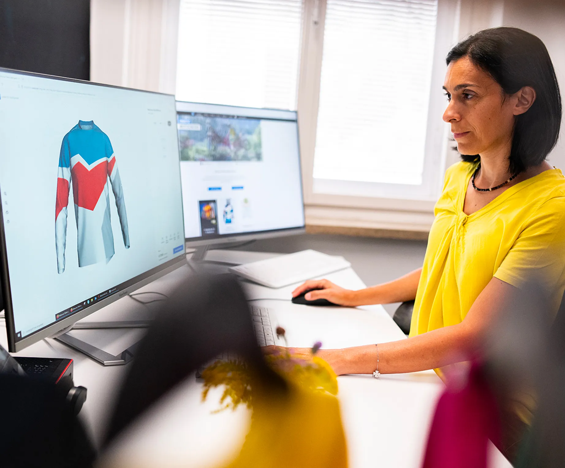 Eine Frau in einem gelben Oberteil sitzt an einem Schreibtisch und entwirft auf einem Computerbildschirm ein maßgeschneidertes Sporttrikot mit blauen, roten und weißen Farbflächen.