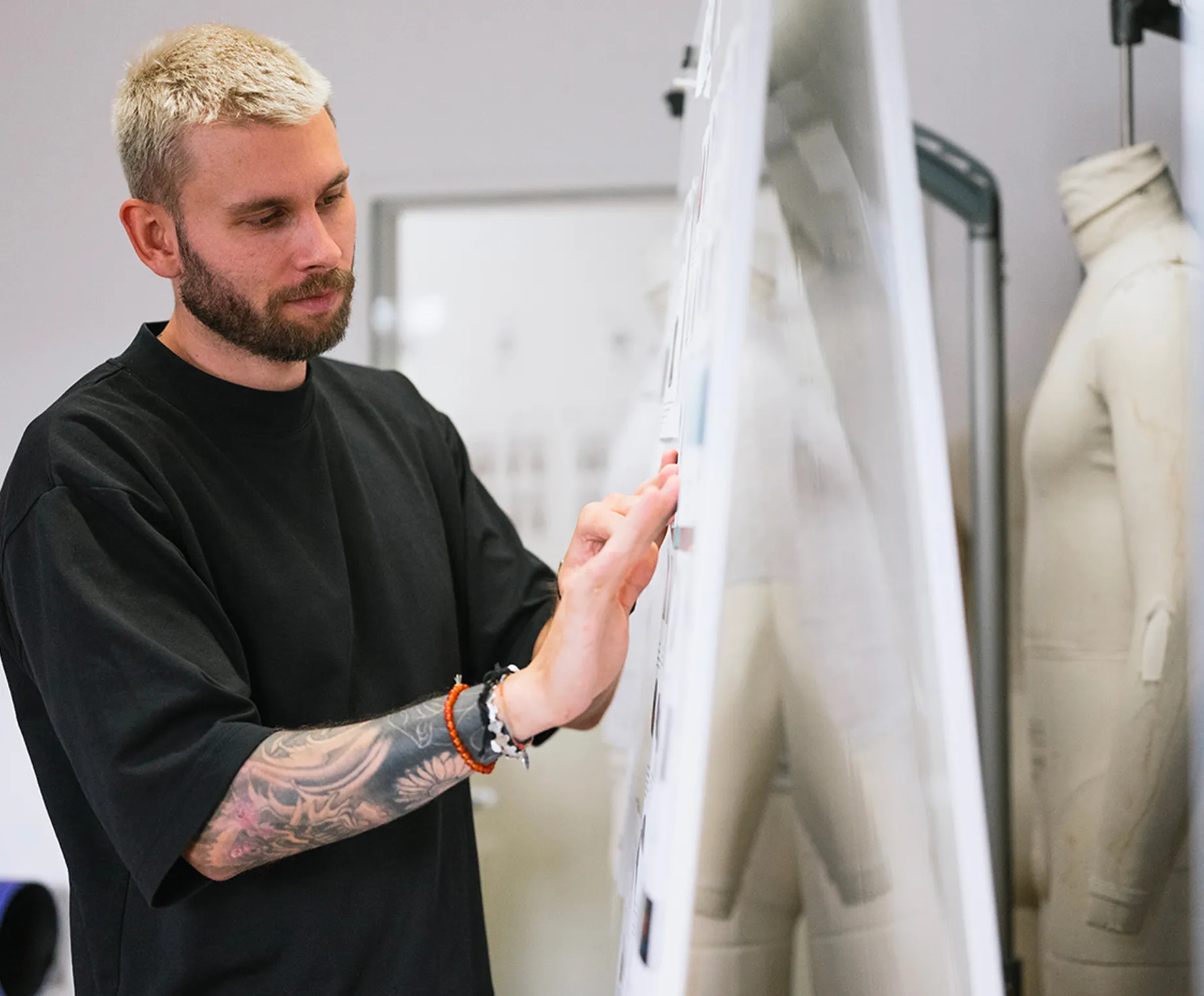 Ein Mann mit blondem Haar und einem Tattoo am Arm betrachtet konzentriert Entwürfe auf einer Pinnwand in einem Designstudio, während eine Schneiderpuppe im Hintergrund steht.