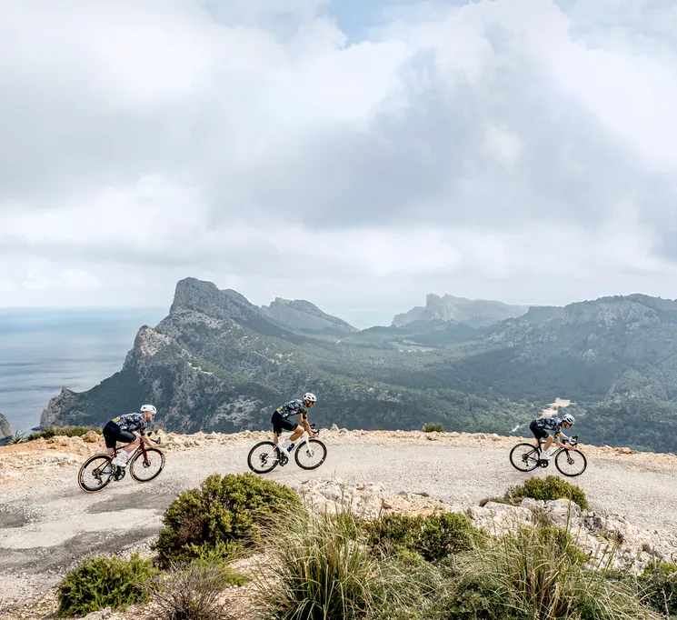 Drei Radfahrer in sportlicher Kleidung fahren auf einer Straße entlang einer Küstenlandschaft mit Bergen und Meer im Hintergrund.